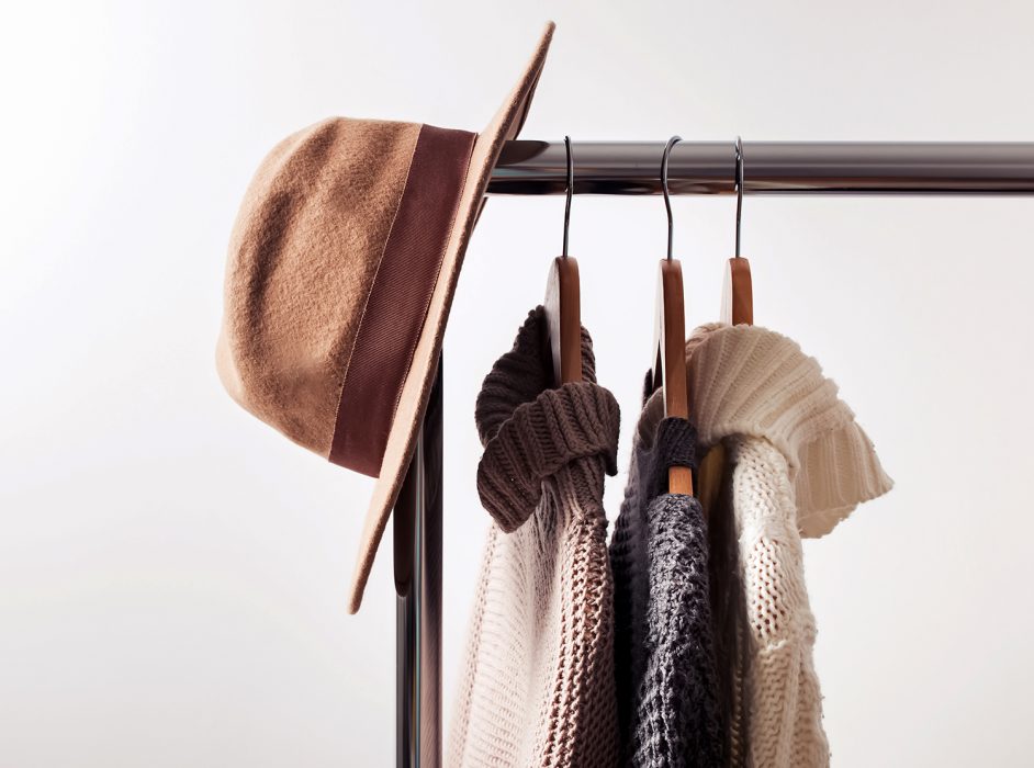 Knitted sweaters on hangers and felt hat Knitted sweaters on hangers and felt hat on the white wall background. Autumn or winter wardrobe.